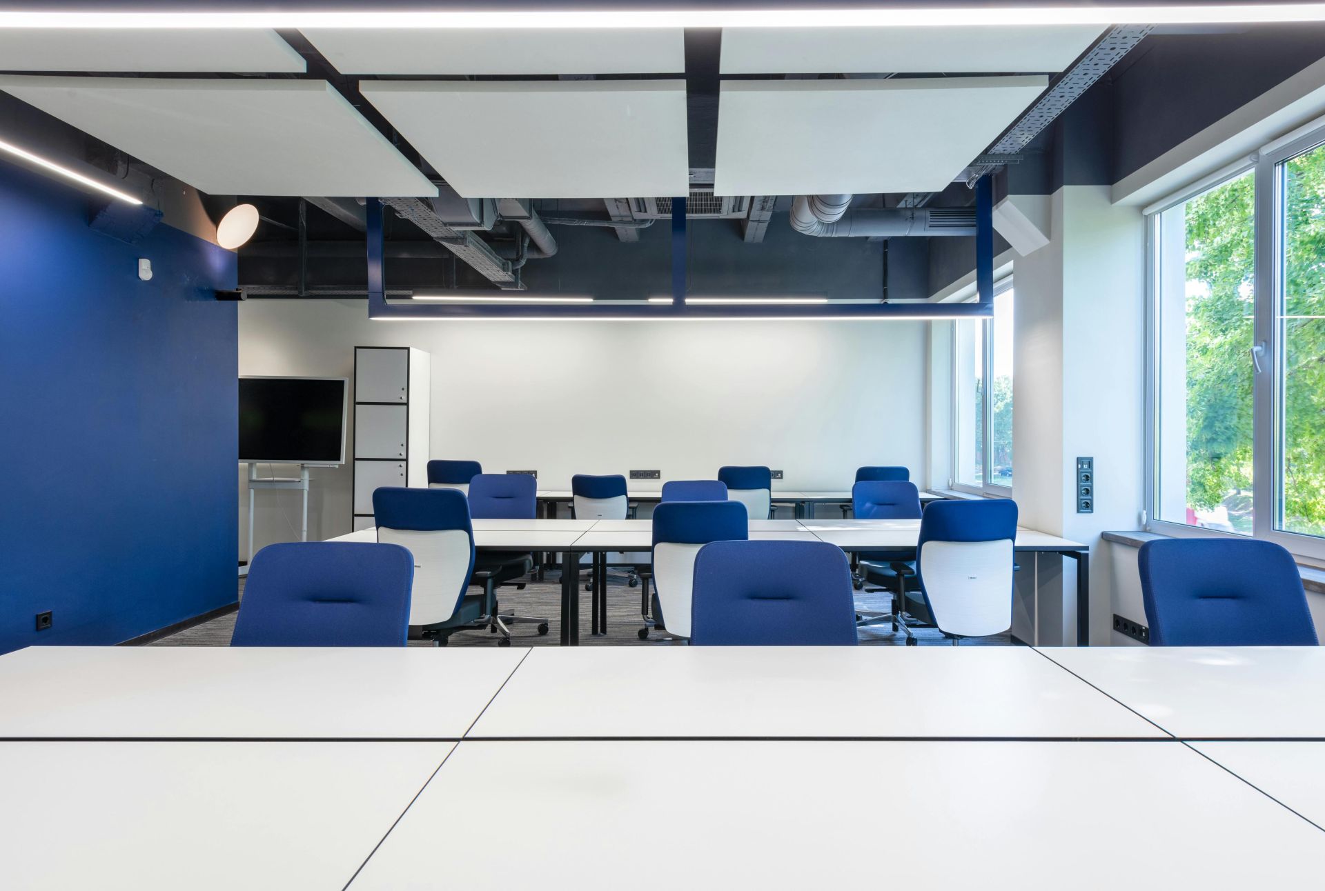 Various white desks with blue chairs placed in modern spacious workspace with big window and monitor in contemporary business center